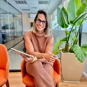 Dra. Elyssa Araújo, advogada especialista em direito do trabalho, sorrindo de forma acolhedora com um tablet em mãos. Ela está sentada em um escritório corporativo moderno em Niterói, pronta para o atendimento consultivo.