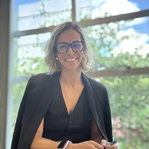 Dra. Elyssa Araújo sorrindo, vestindo um blazer escuro sobre os ombros e óculos, sentada próxima a uma janela com luz natural. A foto reflete sua sólida qualificação acadêmica e expertise em compliance e direito digital.
